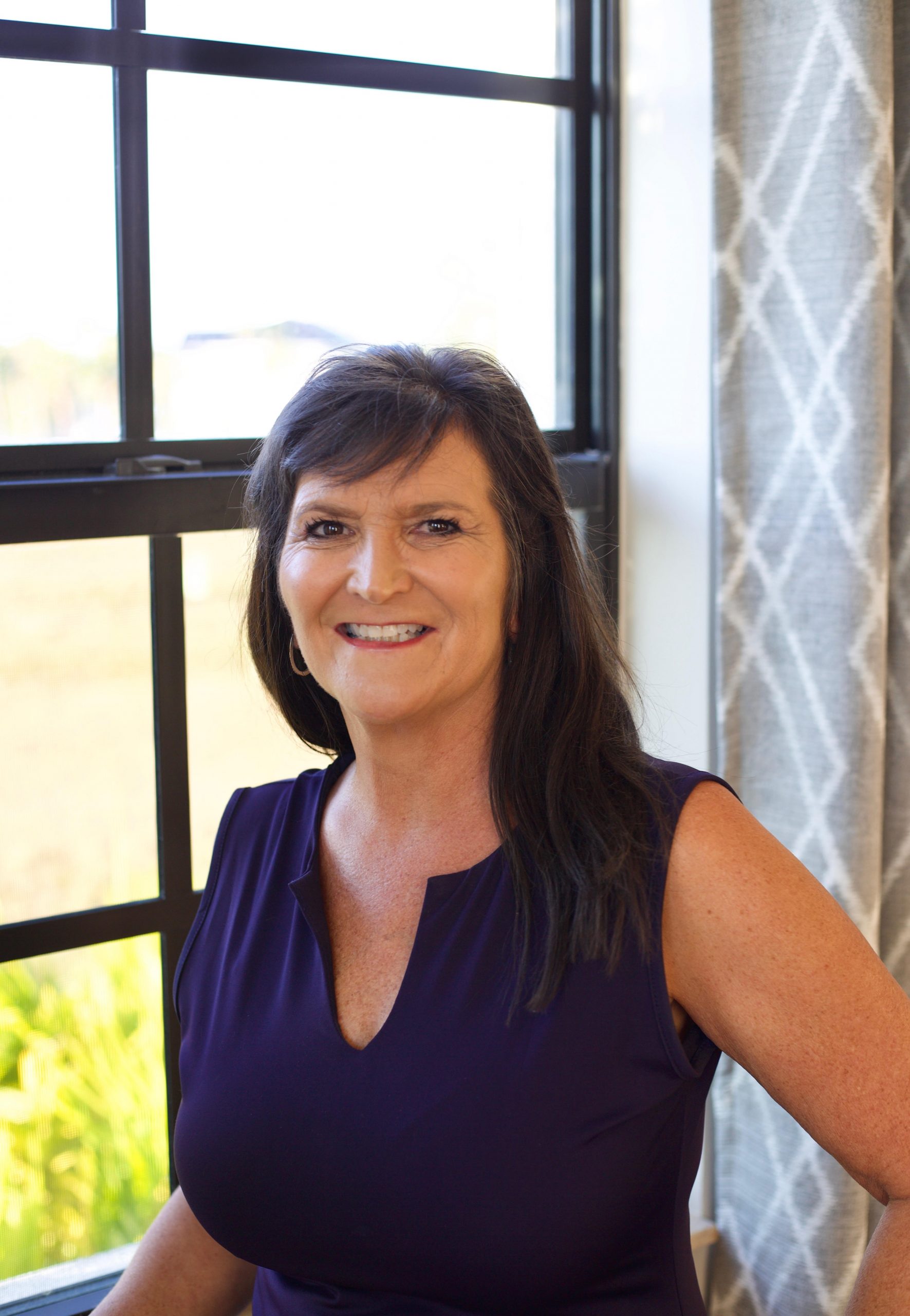 Cheryl Hayek smiling in front of sunny window with a decorative curtain